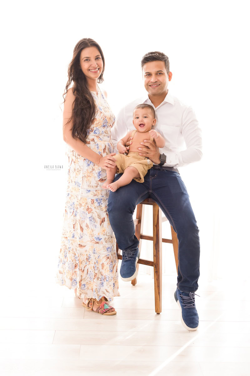 10 months boy posing seated on dad's lap, on a stool with mom standing, against a serene white backdrop, captured by Anega Bawa Family Photographer in Gurgaon (Delhi NCR).