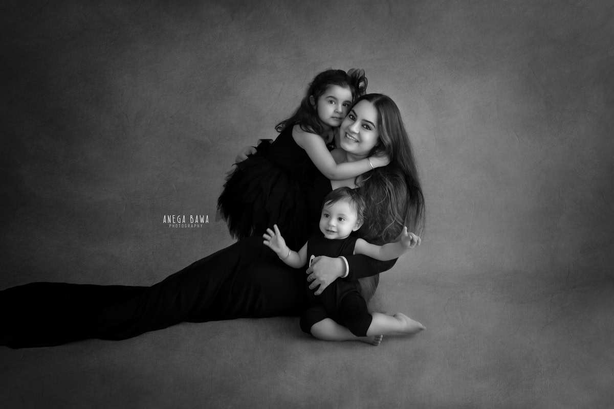 10-month-old boy posing with his elder sister and mom, all dressed in black, against a black and white backdrop, captured in a stylish family portrait by Anega Bawa, Gurgaon (Delhi, NCR)