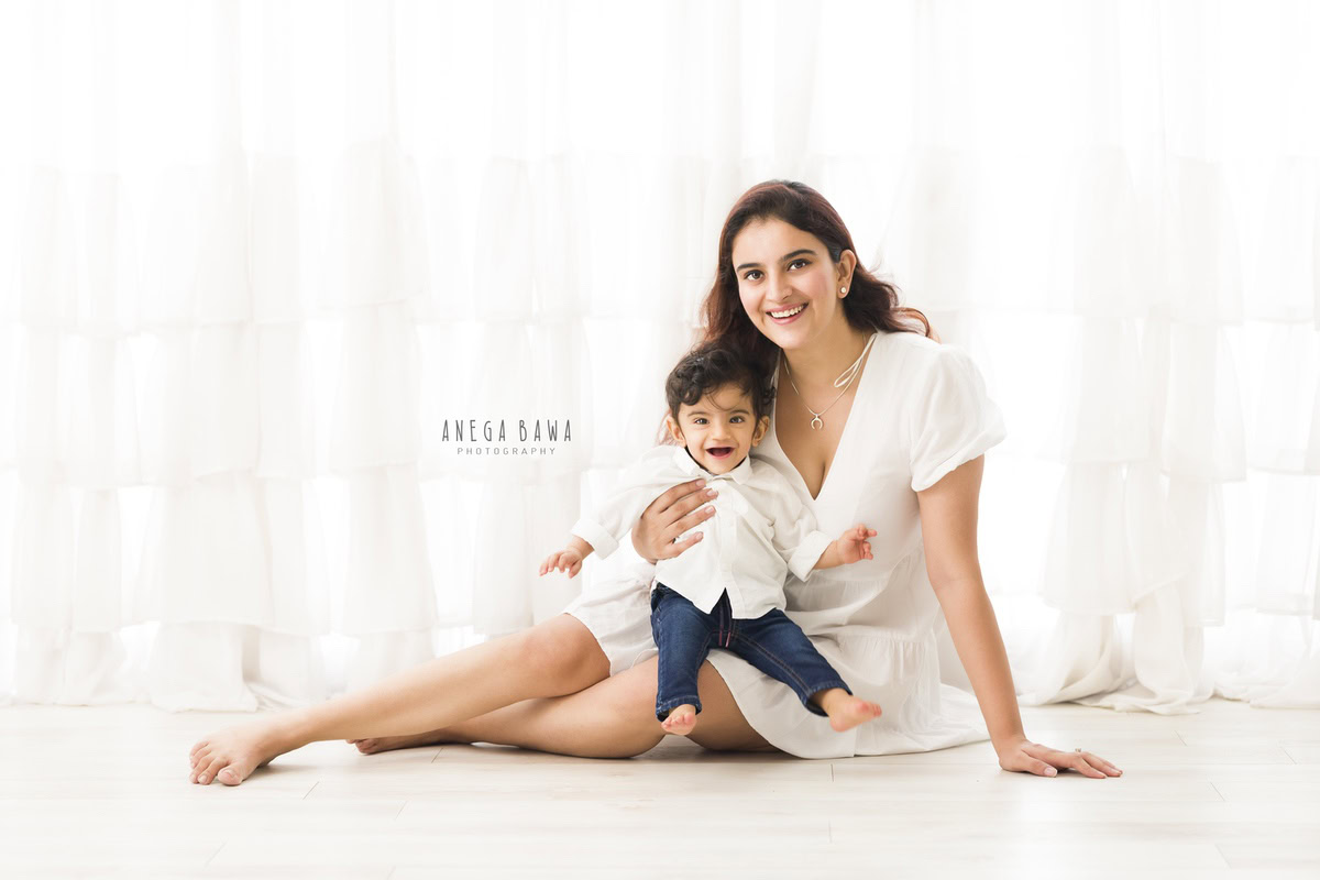 10-months boy seated on mom's lap, smiling cutely against a white backdrop, captured by Anega Bawa Family Photographer in Gurgaon (Delhi NCR).