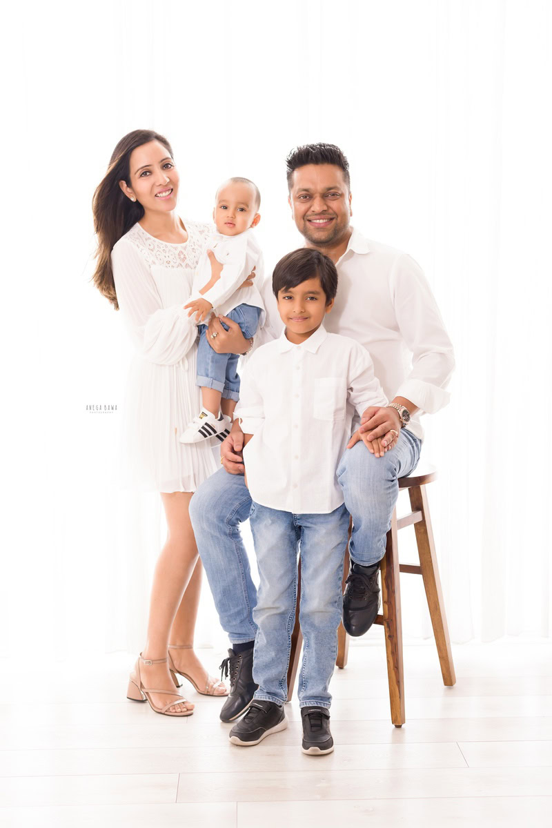 10-months girl in mom's arms, with elder brother and dad, all dressed in white shirts, against a white backdrop, captured by Anega Bawa Family Photographer in Gurgaon (Delhi NCR).