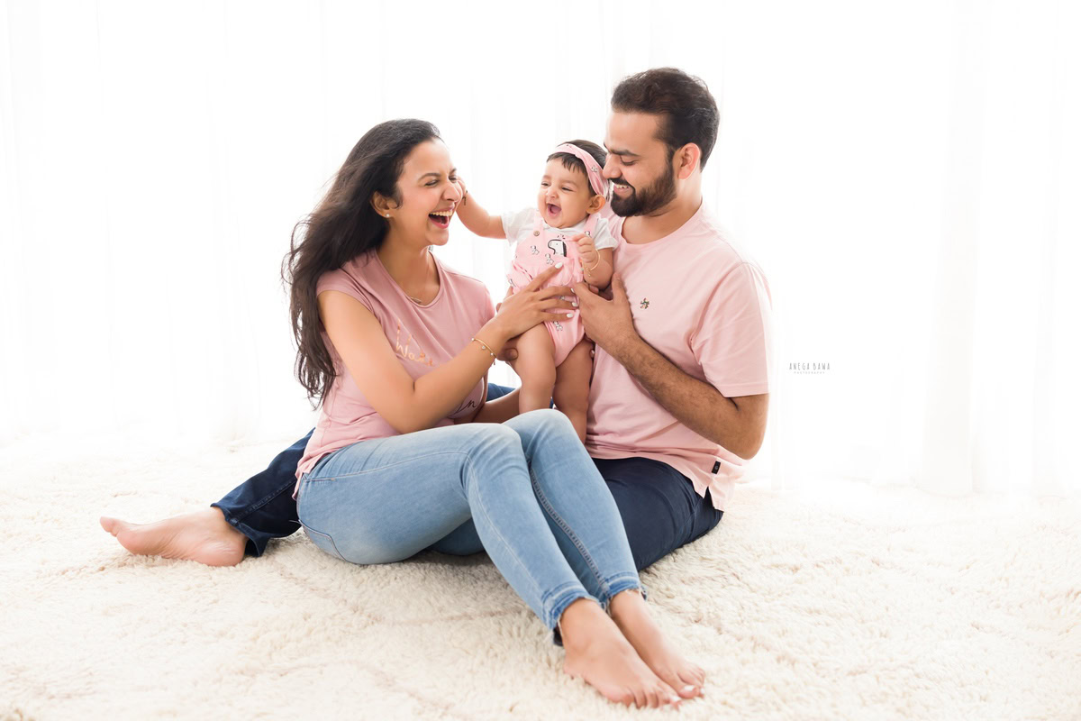 10-months girl playing with mom and dad, seated on a beige rug against a serene white backdrop, captured by Anega Bawa Family Photographer in Gurgaon (Delhi NCR).