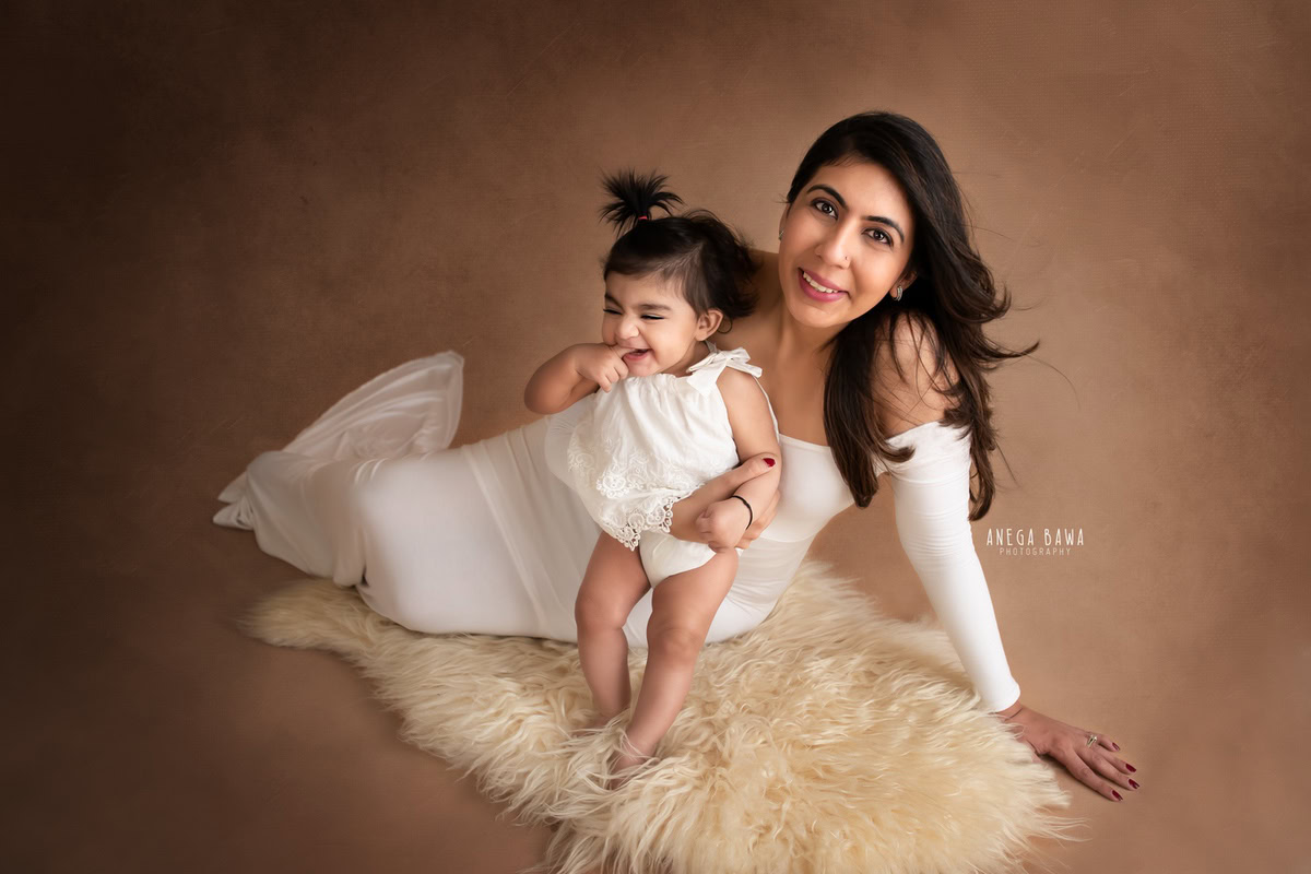 Adorable 10-month-old girl posing with mom, seated on a beige rug, both dressed in white, against an orange peach backdrop, beautifully captured by Anega Bawa in Gurgaon (Delhi, NCR)