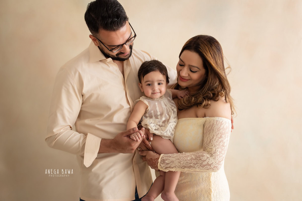 10-month-old girl posing with mom and dad against a soft beige backdrop, captured beautifully by Anega Bawa in Gurgaon (Delhi, NCR)
