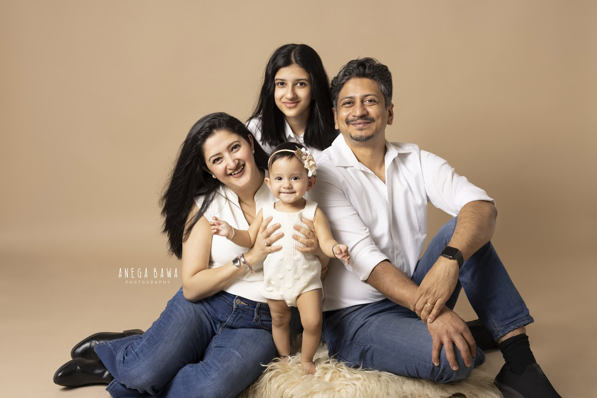 10-month-old girl posing with mom, dad, and elder sister seated on a beige rug against a light brown backdrop, all wearing white. Captured by Anega Bawa Photography, Gurgaon (Delhi, NCR).