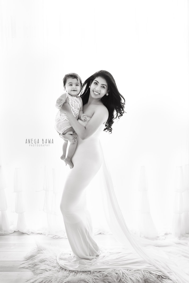 10-month-old girl posing with mom, wearing a gown, standing on a beige rug against a white backdrop, captured by Anega Bawa family photographer Gurgaon (Delhi NCR).