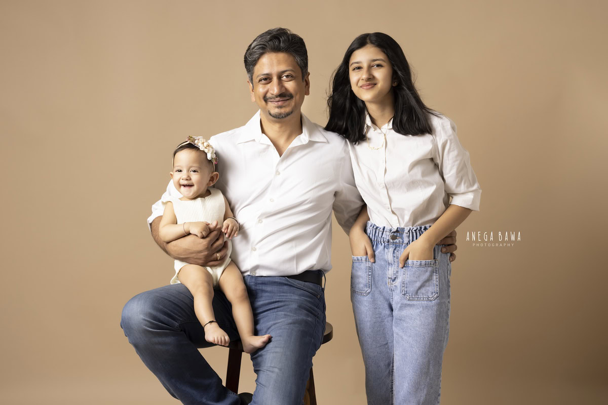 10-months girl seated on dad's lap, all wearing white, against a light brown backdrop, captured by Anega Bawa Family Photographer in Gurgaon (Delhi NCR).
