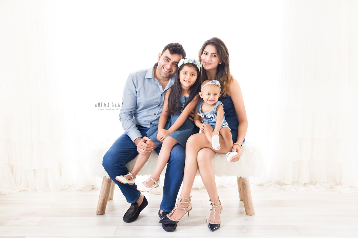 10-month-old girl seated on mom's lap, posing with dad and elder sister against a clean white backdrop. Captured by Anega Bawa Photography, Gurgaon (Delhi, NCR).