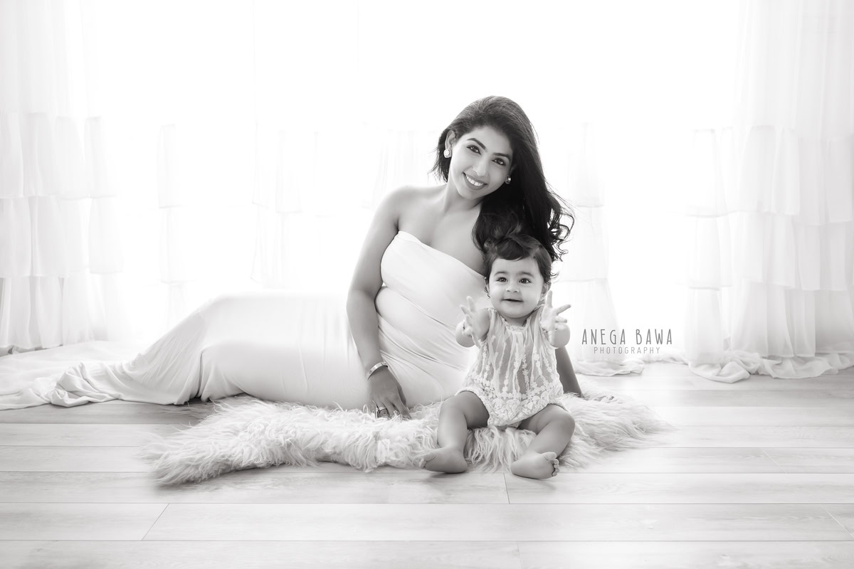 10-month-old girl seated with mom, smiling against a white backdrop with a beige rug, captured by Anega Bawa Photography for a family photoshoot in Gurgaon (Delhi NCR).