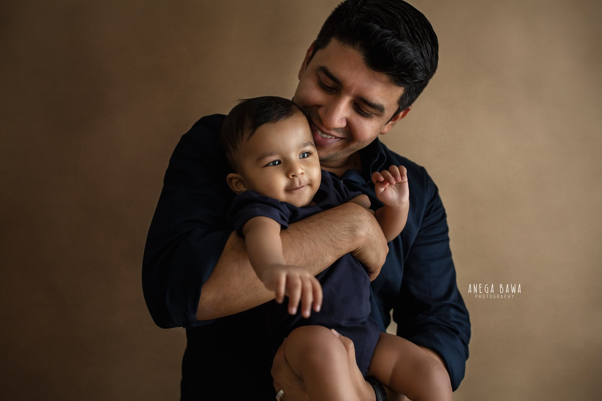 10-month-old son in dad's arms against a light brown backdrop, striking an adorable kissing pose. Captured by Anega Bawa, family photographer in Gurgaon (Delhi NCR).