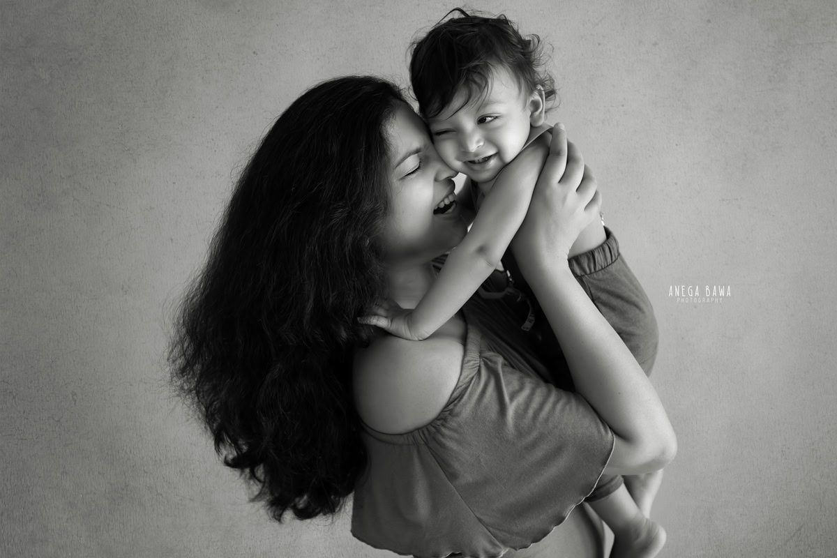 10-month-old son in mom's arms, posing cutely against a light brown backdrop, captured by Anega Bawa family photographer Gurgaon (Delhi NCR).