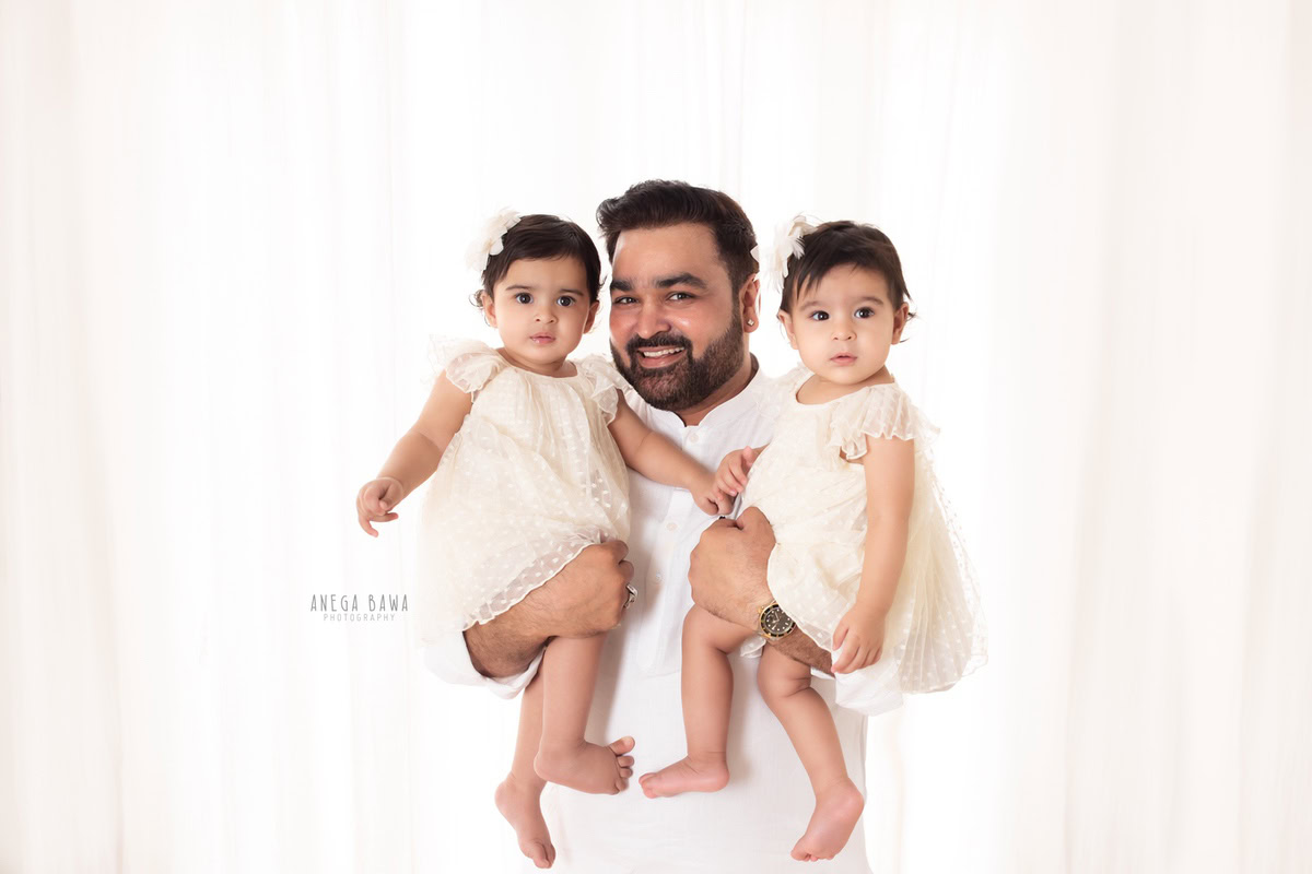 10-month-old twin girls posing with dad against a white backdrop, all dressed in white, captured beautifully by Anega Bawa in Gurgaon (Delhi, NCR)