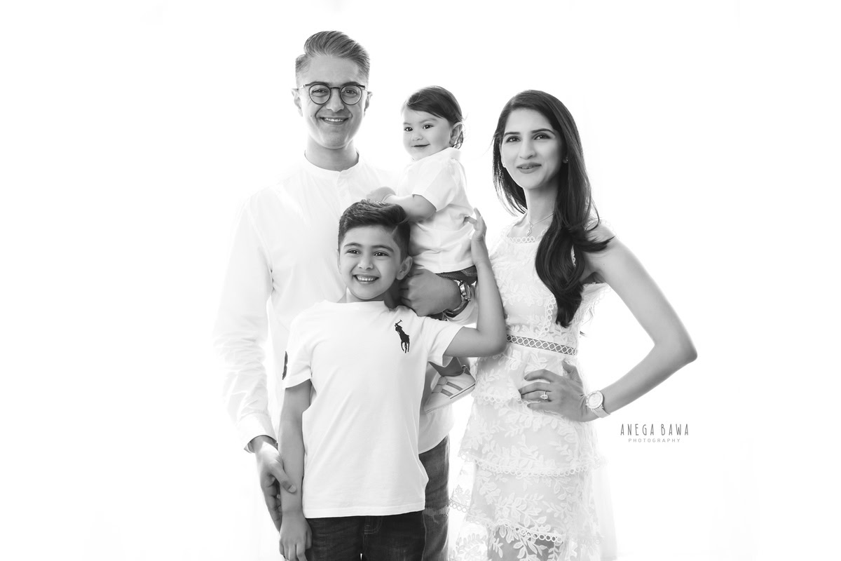 11-month-old boy in dad's arms, posing with elder brother and mom in a family pose against a white backdrop, all dressed in white, captured by Anega Bawa in Gurgaon (Delhi, NCR)