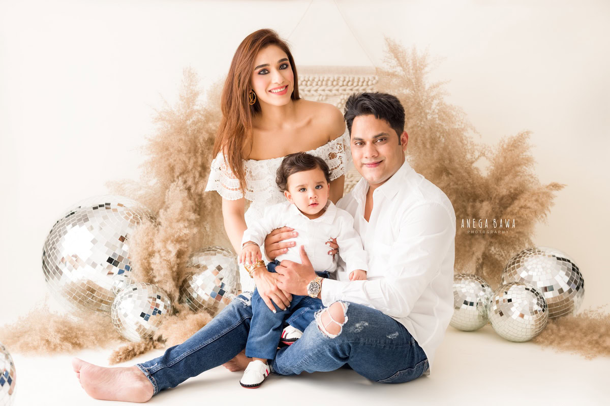 11-month-old boy seated on dad's lap with mom, surrounded by bushes and disco balls, all wearing white against a white backdrop. Captured by Anega Bawa, family photographer in Gurgaon (Delhi NCR).