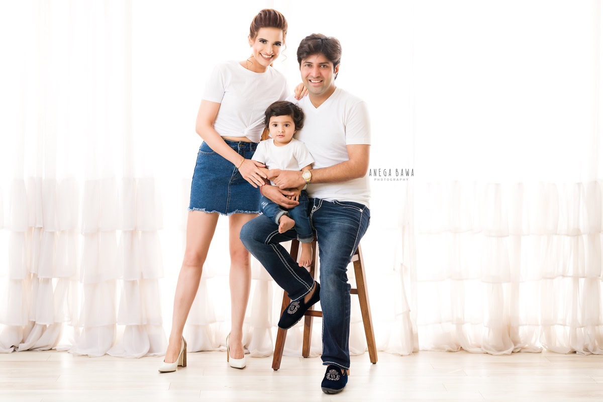 11-month-old boy seated on dad's lap, mom standing, all wearing white tees and blue denim against a white backdrop, captured by Anega Bawa family photographer Gurgaon (Delhi NCR).
