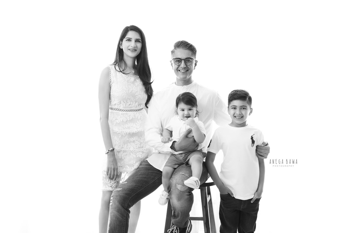 11-month-old boy seated on dad's lap, posing with mom and elder brother, all dressed in white against a clean white backdrop. Captured by Anega Bawa, family photographer in Gurgaon (Delhi NCR).