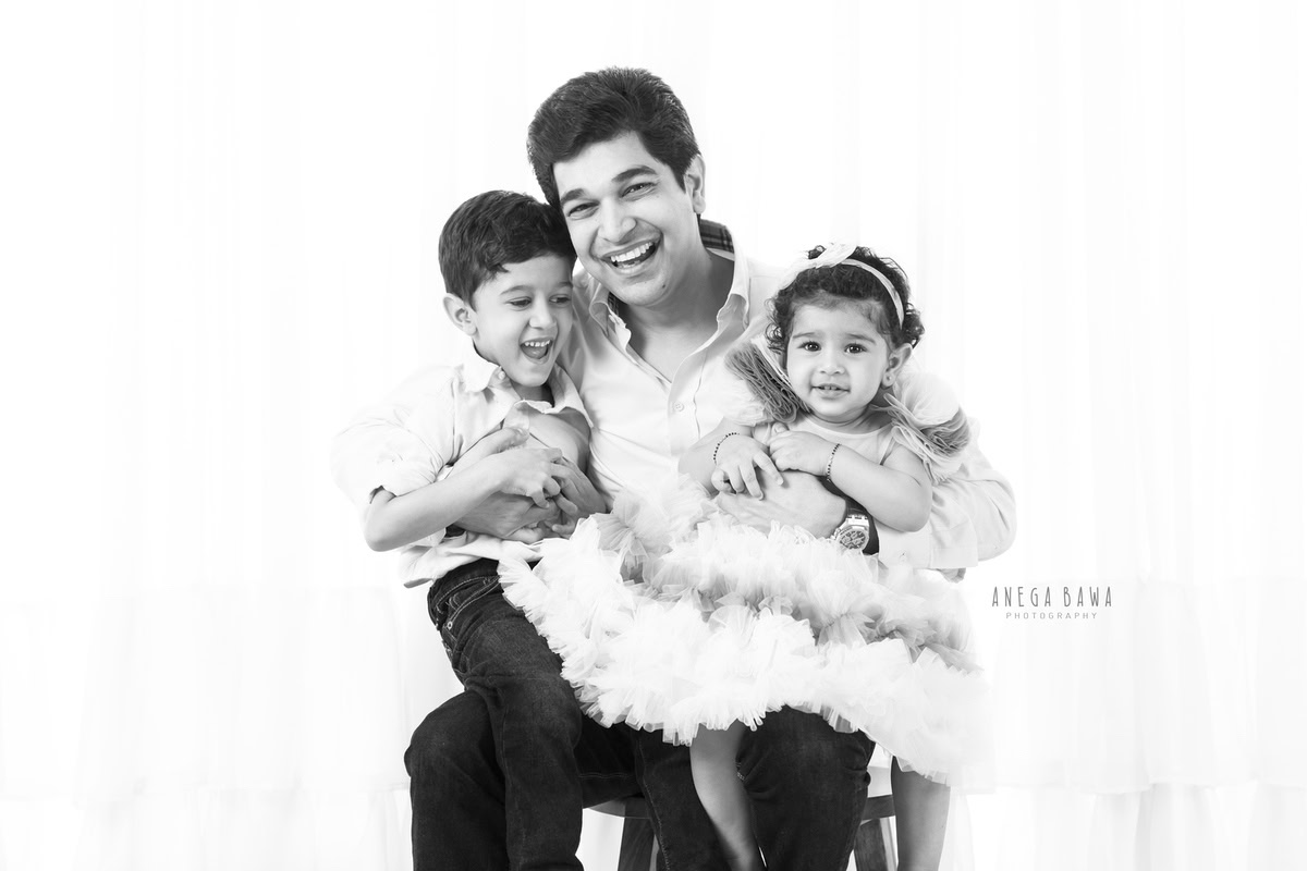 11-month-old girl and elder brother seated on dad's lap, smiling against a white backdrop, captured by Anega Bawa in Gurgaon (Delhi, NCR)
