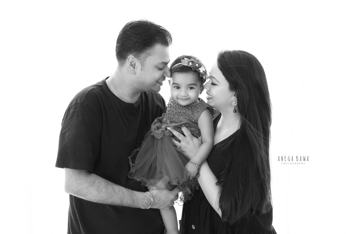 11-month-old girl posing with mom and dad against a white backdrop, beautifully captured by Anega Bawa Photography for a family photoshoot in Gurgaon (Delhi NCR).