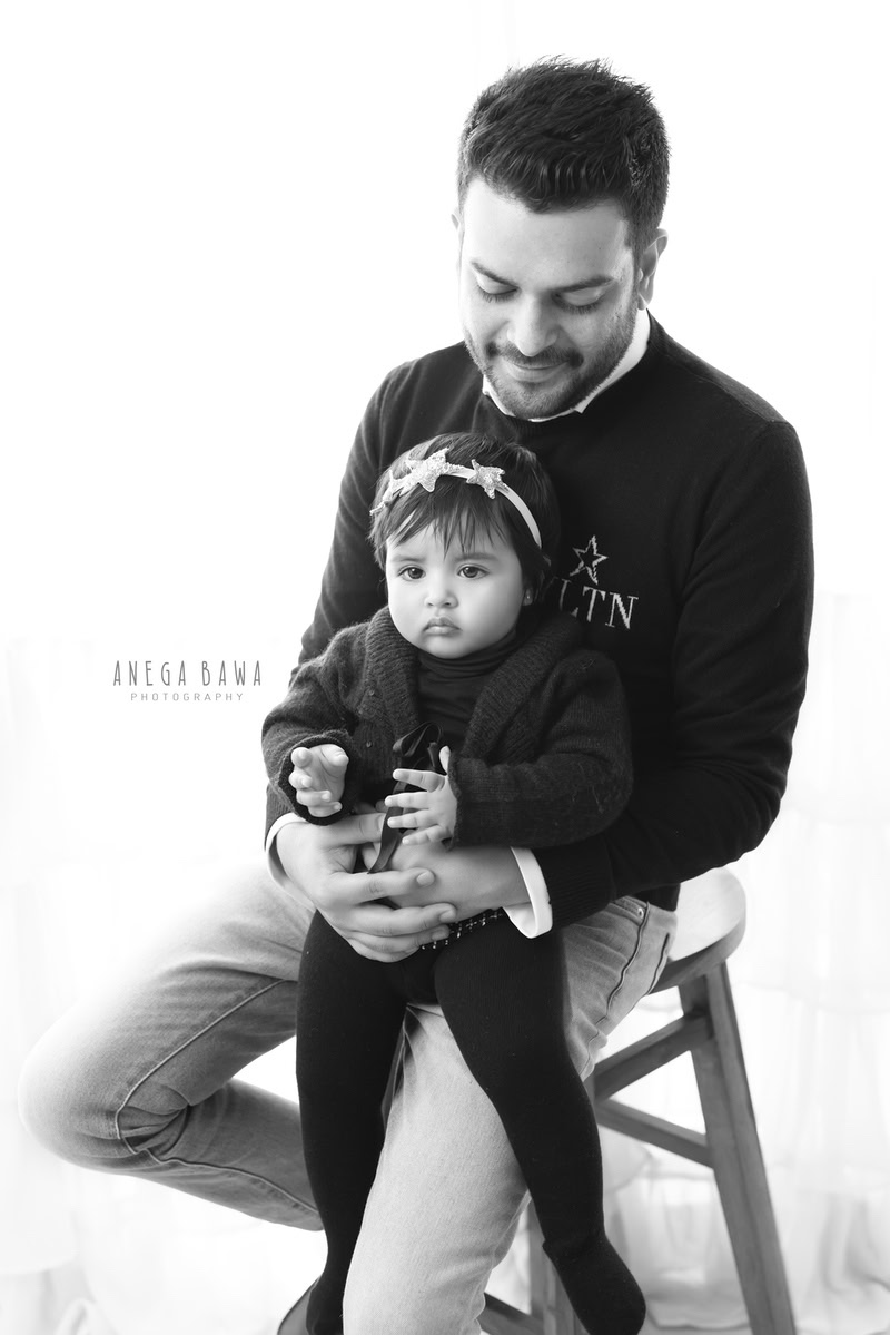 11-month-old girl seated on dad's lap against a clean white backdrop. Captured by Anega Bawa Photography, Gurgaon (Delhi NCR).