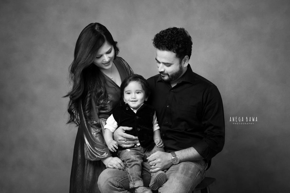 12-month-old boy sitting on dad's lap with mom posing cutely against a black and white backdrop, beautifully captured by Anega Bawa Photography for a family photoshoot in Gurgaon (Delhi NCR).