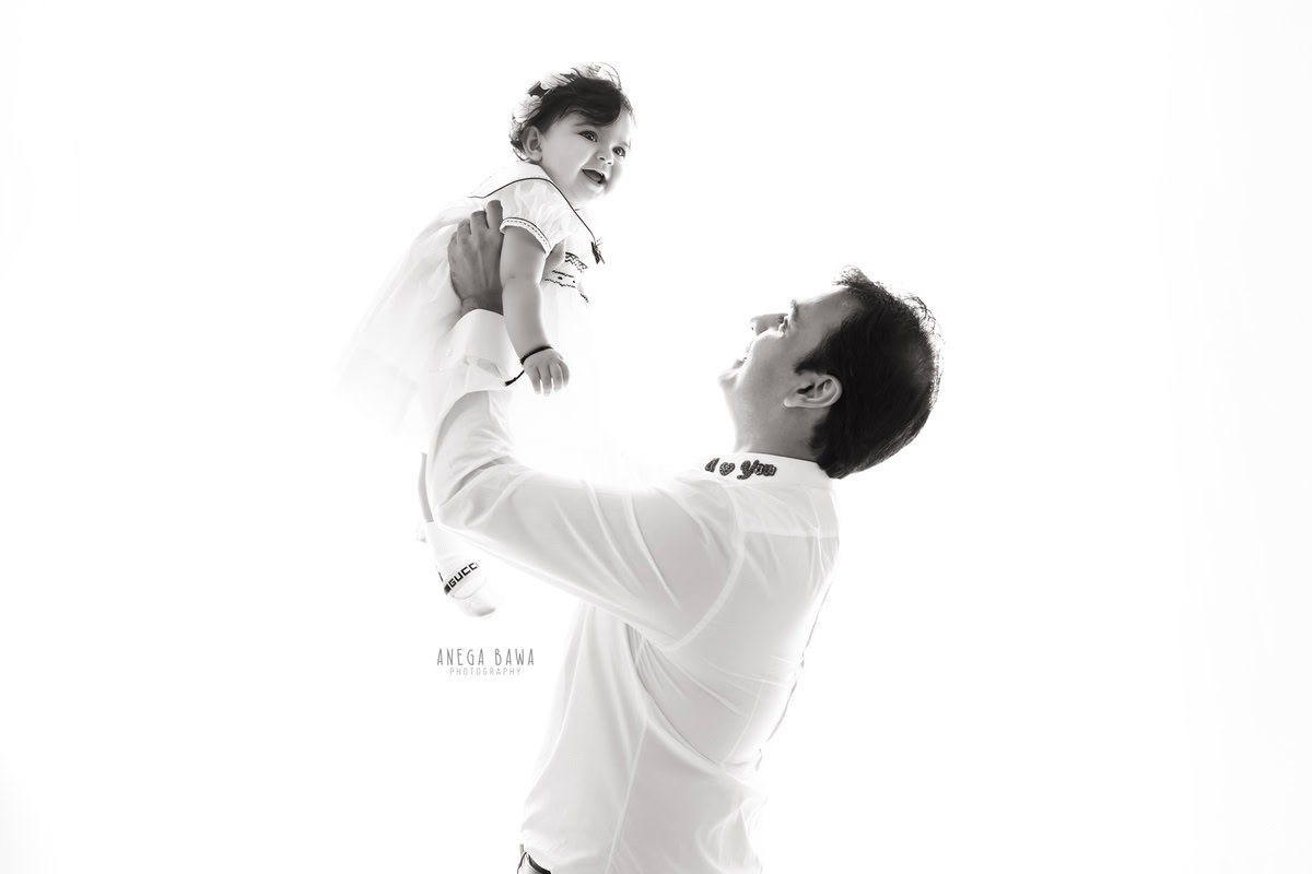 12-month-old girl in dad's arms wearing white against a white backdrop, beautifully captured by Anega Bawa Photography for a family photoshoot in Gurgaon (Delhi NCR).