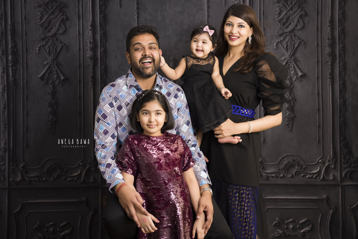 12-month-old girl in mom's arms, playfully interacting with dad's ears, with elder sister in a family pose against a black backdrop. Captured by Anega Bawa Photography, Gurgaon (Delhi NCR).