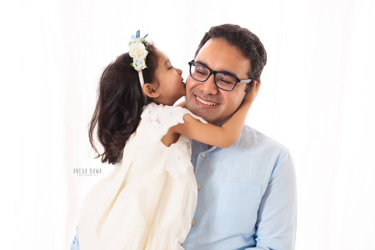 12-month-old girl posing cutely with dad in a kissing pose against a white backdrop, beautifully captured by Anega Bawa Photography for a family photoshoot in Gurgaon (Delhi NCR).
