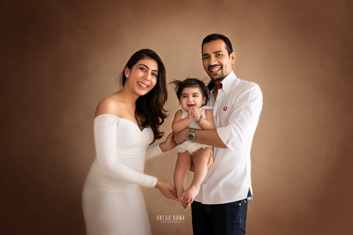 12-month-old girl posing cutely with mom and dad against a peach-orange backdrop, all wearing white, beautifully captured by Anega Bawa Photography for a family photoshoot in Gurgaon (Delhi NCR).