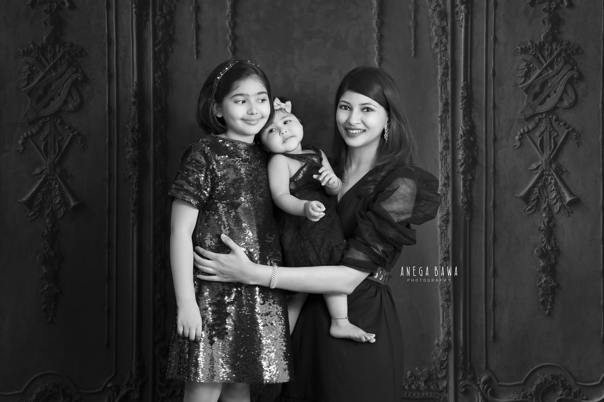 12-month-old girl posing with mom and elder sister against a dramatic black backdrop. Captured by Anega Bawa Photography, Gurgaon (Delhi NCR).