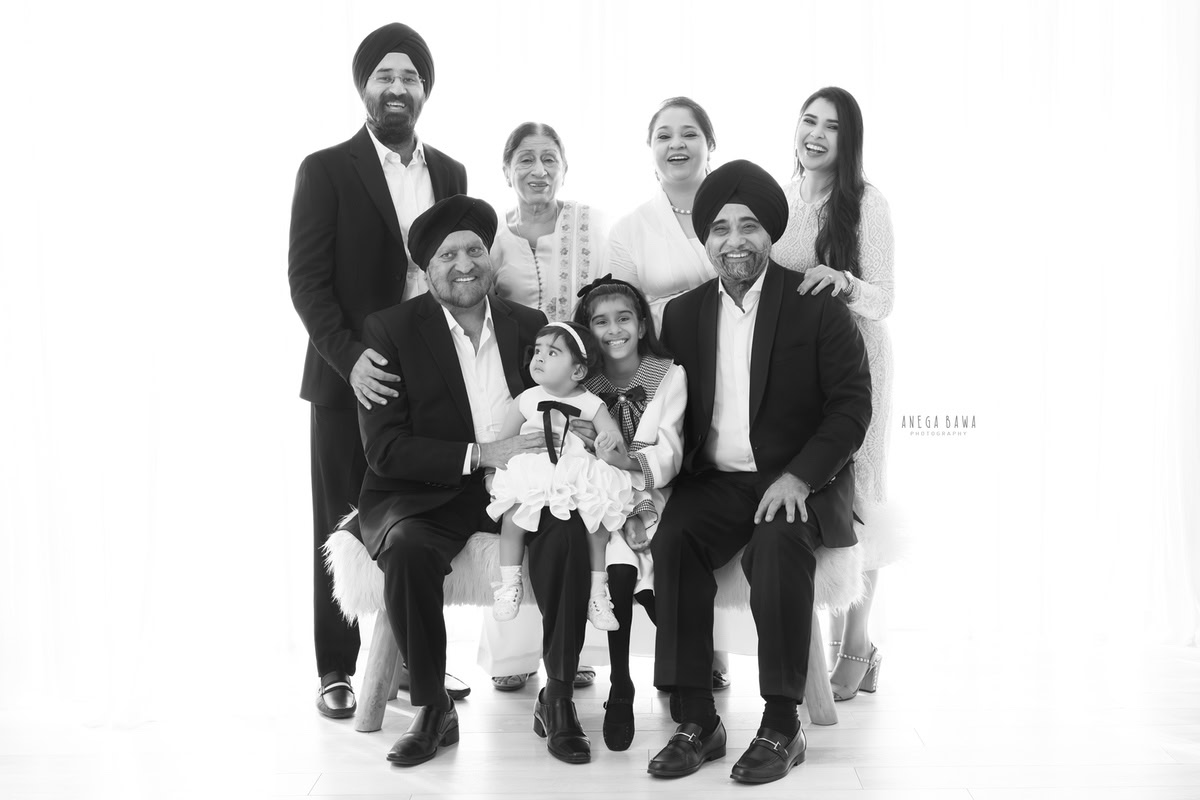 12-month-old girl seated on her grandfather's lap, posing with the whole family against a white backdrop in a smiling pose, beautifully captured by Anega Bawa Photography for a family photoshoot in Gurgaon (Delhi NCR).