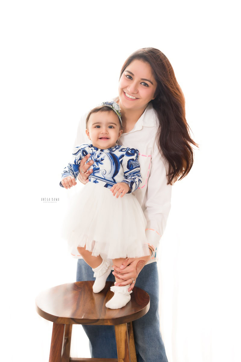12-month-old girl standing on a wooden stool against a clean white backdrop, posing with mom. Captured by Anega Bawa Photography, Gurgaon (Delhi NCR).