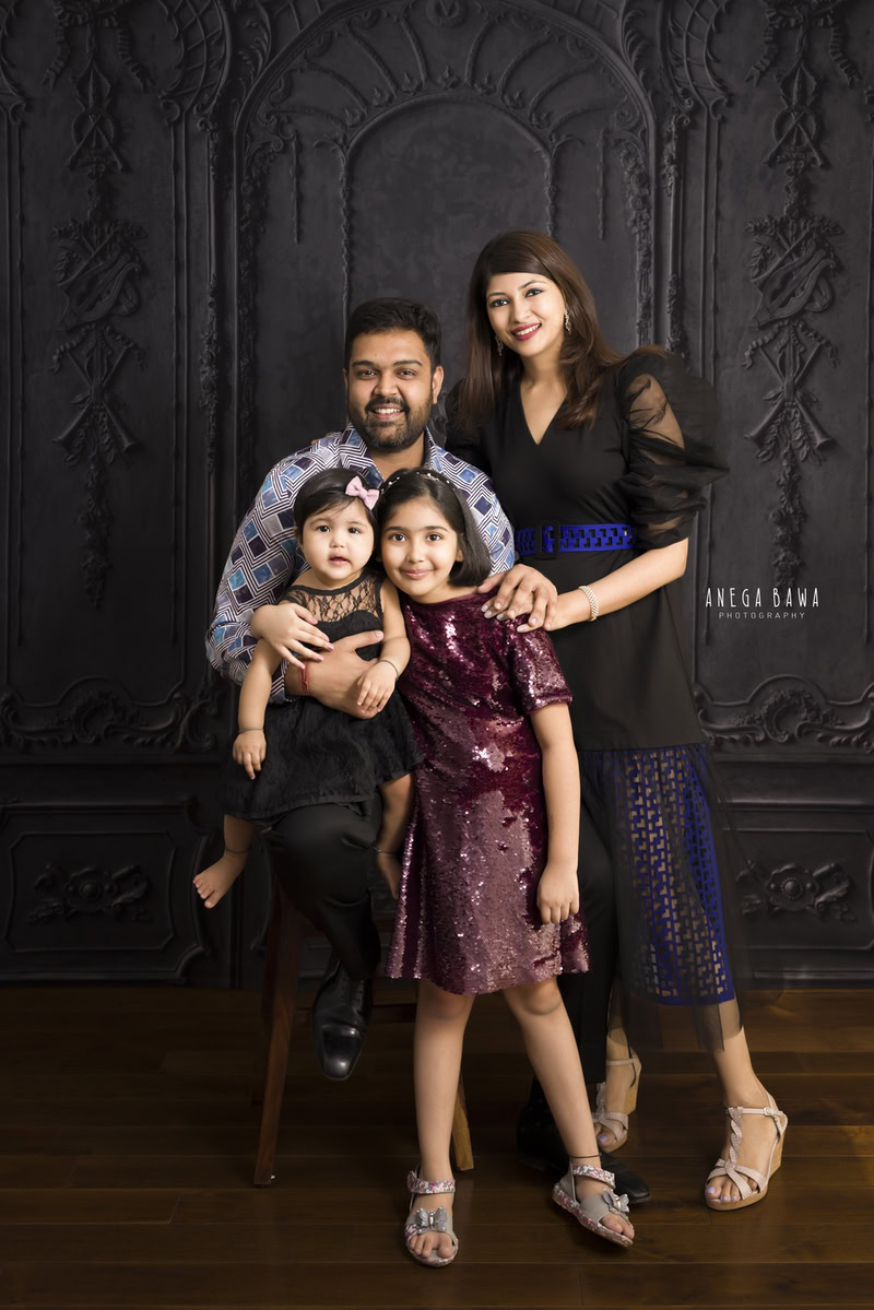 12-month-old girl wearing a cute black dress, posing with mom, dad, and elder sister against a black backdrop, beautifully captured by Anega Bawa Photography for a family photoshoot in Gurgaon (Delhi NCR).