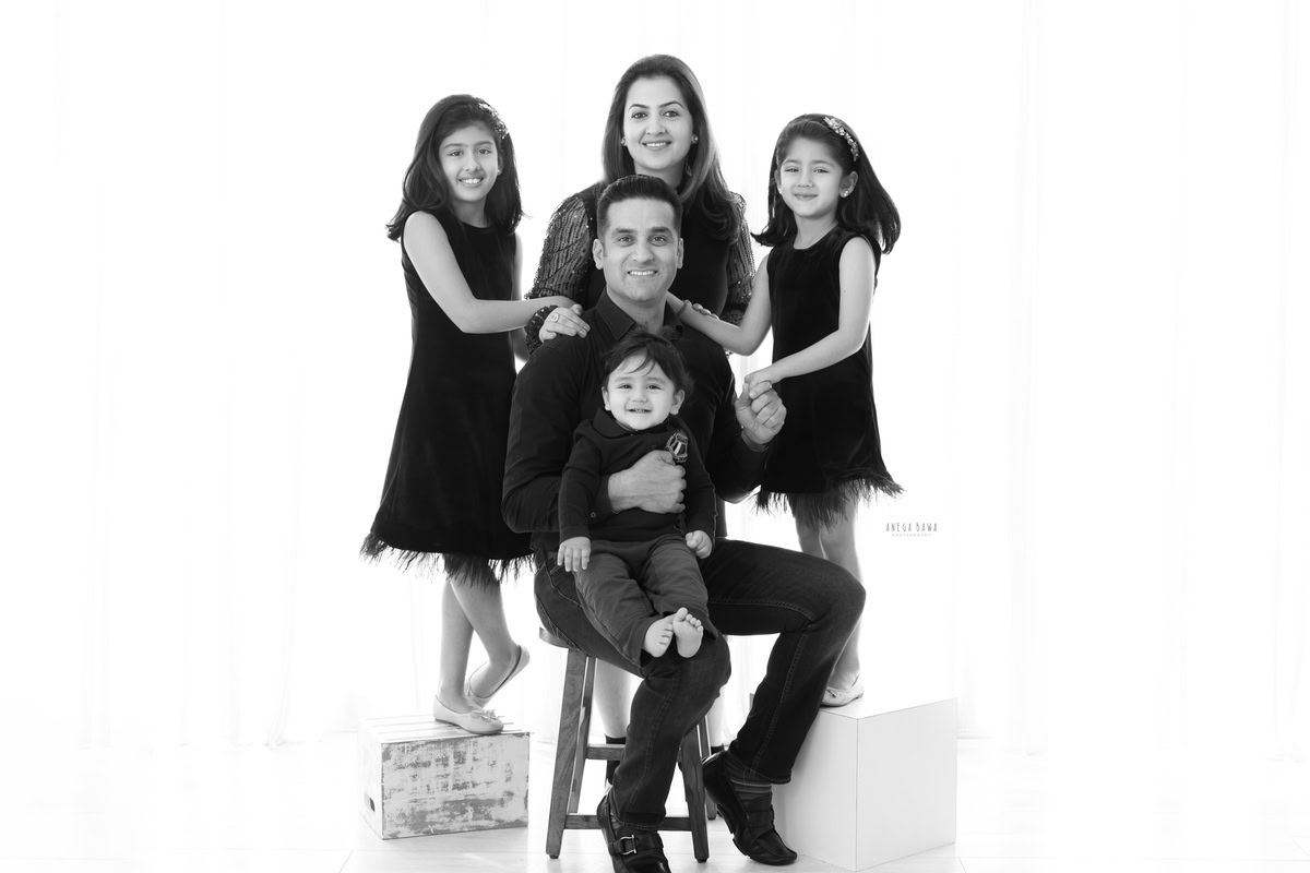 14-month-old boy seating on dad's lap, posing with mom and twin sisters, all wearing black against a white backdrop. This beautiful family pose was captured by Anega Bawa Photography for a family photoshoot in Gurgaon (Delhi NCR).