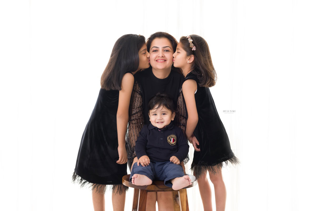 14-month-old boy seated on a stool, posing with mom and twin sisters, all dressed in black against a white backdrop. Captured by Anega Bawa Photography, Gurgaon (Delhi NCR).