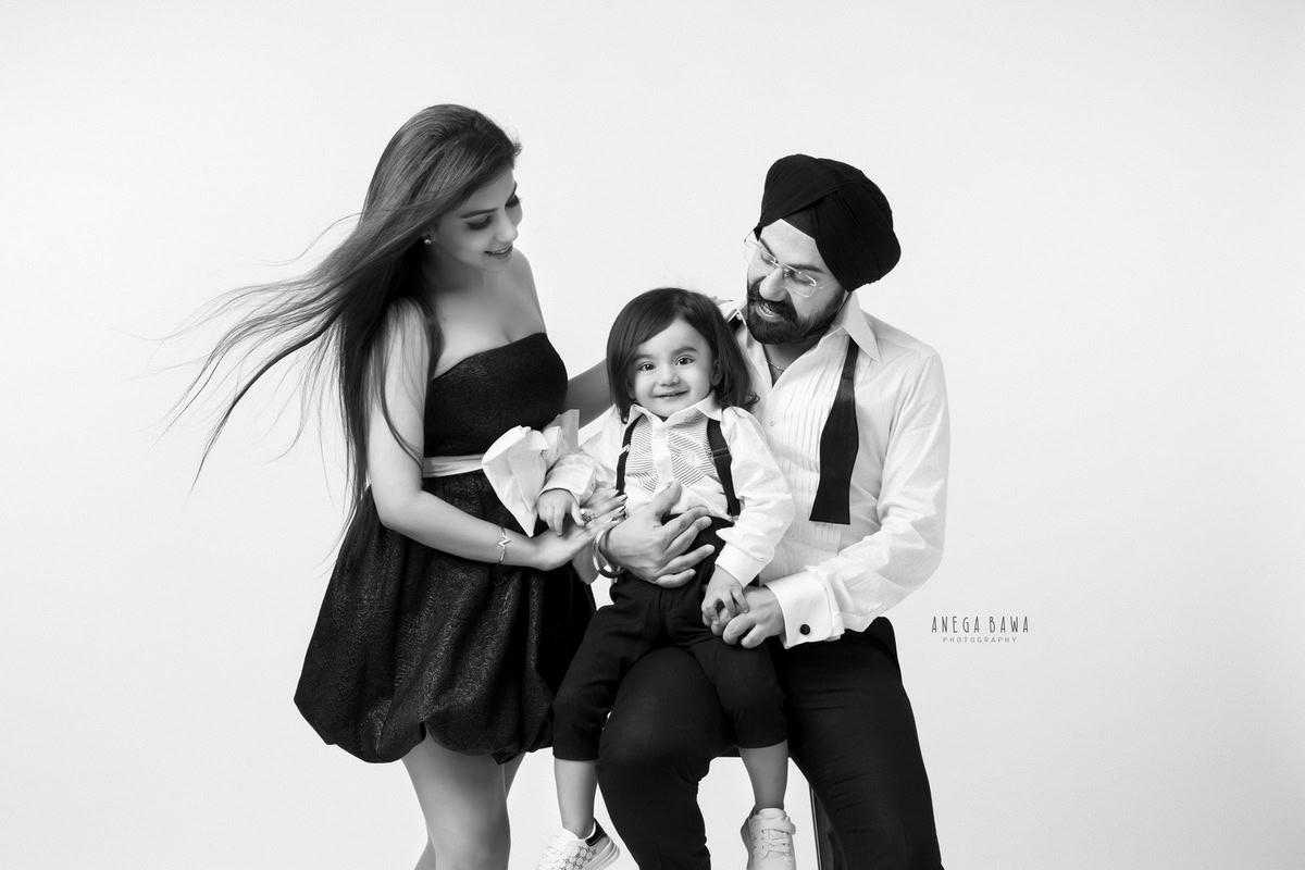 15-month-old boy seated on dad's lap, posing with mom against a clean white backdrop in a black and white pose. Captured by Anega Bawa Photography, Gurgaon (Delhi NCR).