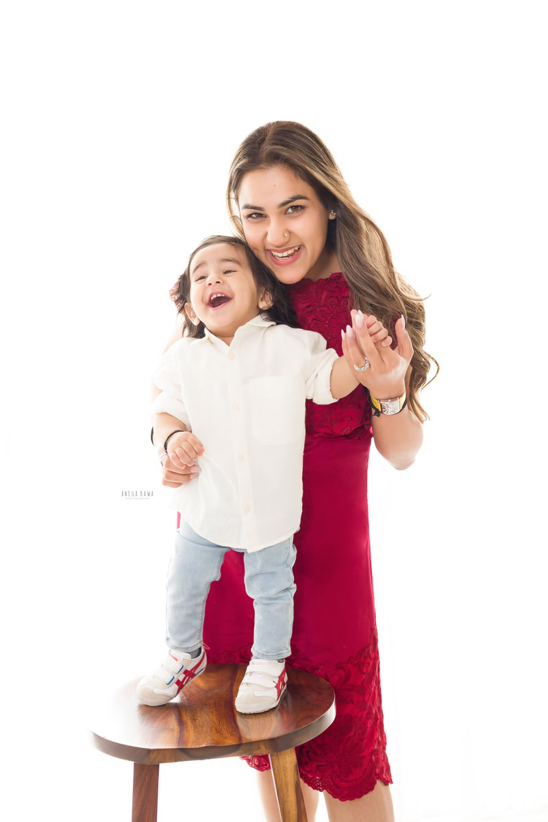 15-month-old boy standing on a wooden stool, laughing in a pose with mom against a white backdrop, captured by Anega Bawa family photographer Gurgaon (Delhi NCR).