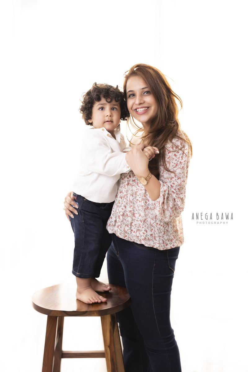 Sweet 15-month-old boy standing on a wooden stool, posing with mom against a serene white backdrop, captured by Anega Bawa Family Photographer Gurgaon (Delhi NCR).
