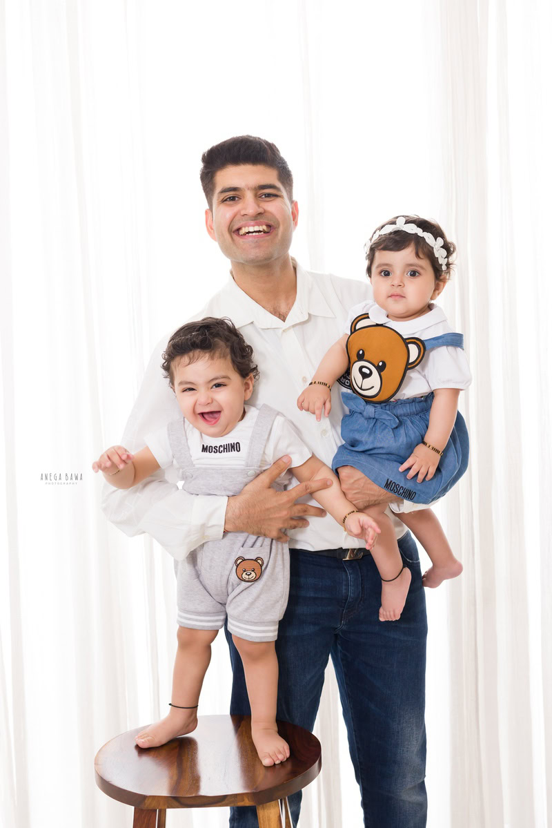 Adorable 15-month-old girl in dad's arms, posing cutely with her elder brother against a white backdrop, captured by Anega Bawa Family Photographer Gurgaon (Delhi NCR).