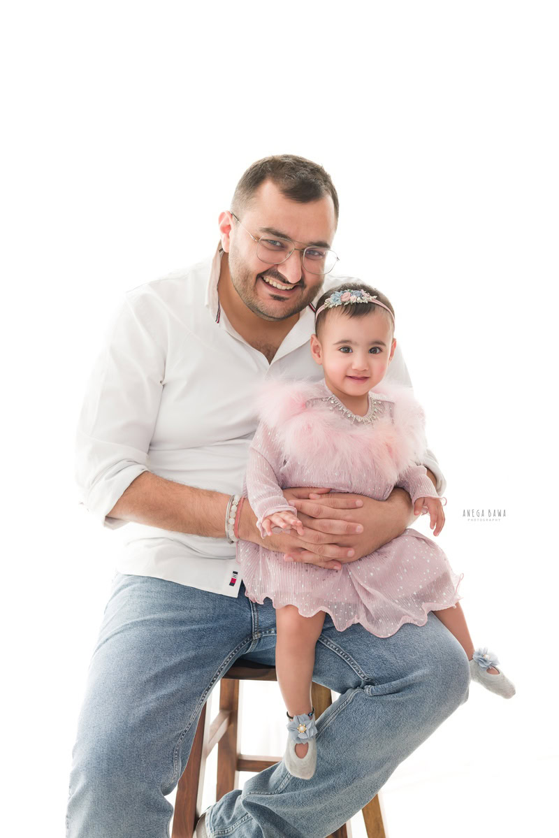 15-months girl seated on dad's lap, wearing a cute headband against a serene white backdrop, captured during a family photoshoot in Gurgaon (Delhi NCR).