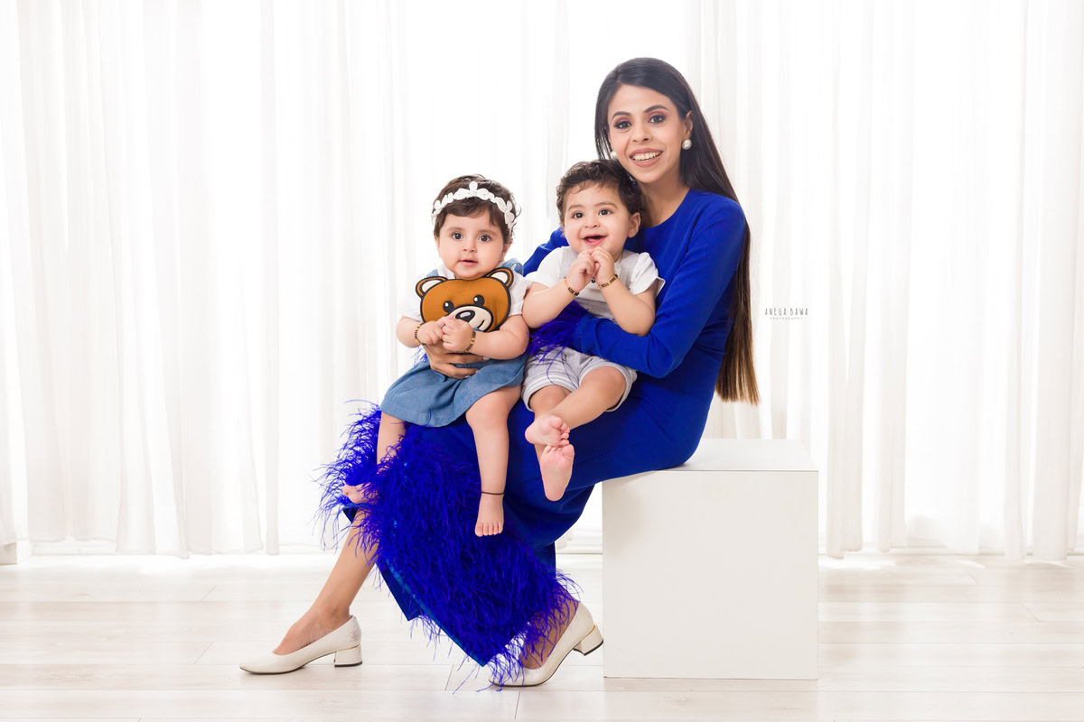 Sweet 15-month-old girl seated on mom's lap with her elder brother against a clean white backdrop, captured by Anega Bawa Family Photographer Gurgaon (Delhi NCR).