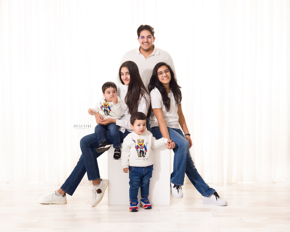 3-year-old boy sitting on mom's lap, posing with his elder sister, dad, and brother in a family pose against a white backdrop. Captured by Anega Bawa Family Photography in Gurgaon (Delhi NCR).