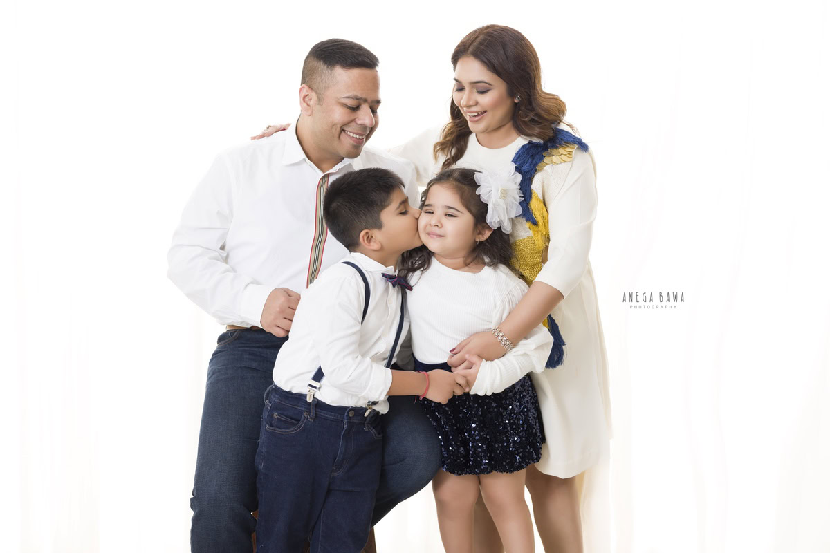 3-year-old girl posing with mom, dad, and elder brother in a sweet kissing pose against a white backdrop. Captured by Anega Bawa Family Photography in Gurgaon (Delhi NCR).