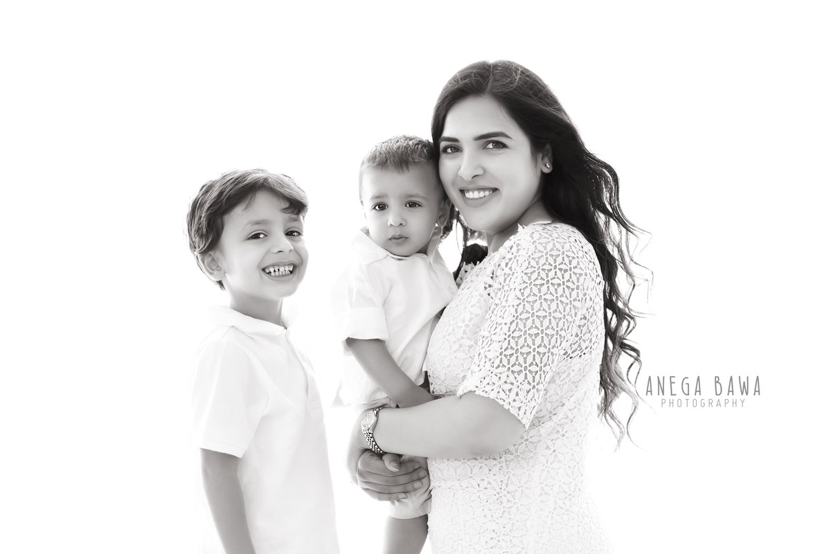 7-month-old son posing with his elder brother and mom, all dressed in white, against a white backdrop, beautifully captured by Anega Bawa Photography for a family photoshoot in Gurgaon (Delhi NCR).