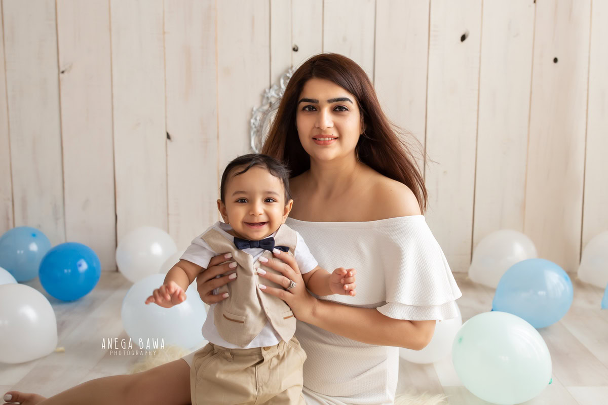 8 months boy posing with mom, seated on floor with blue and white balloons against a beige backdrop, captured by Anega Bawa Family Photographer in Gurgaon (Delhi NCR).