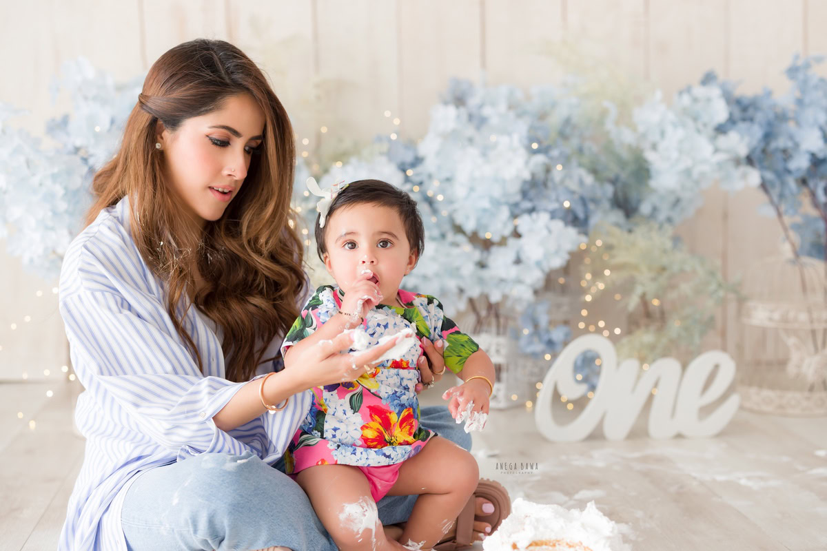 One-year-old girl seated on mom's lap surrounded by blue flowers and fairy lights against a white backdrop, photographed by Anega Bawa in Gurgaon (Delhi, NCR)