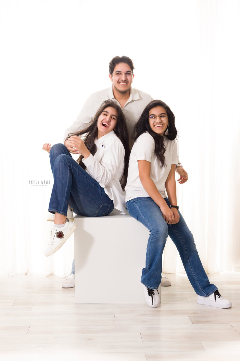 Mom, dad, and daughter seated on a white stool in a smiling pose against a white backdrop, captured by Anega Bawa family photographer Gurgaon (Delhi NCR).