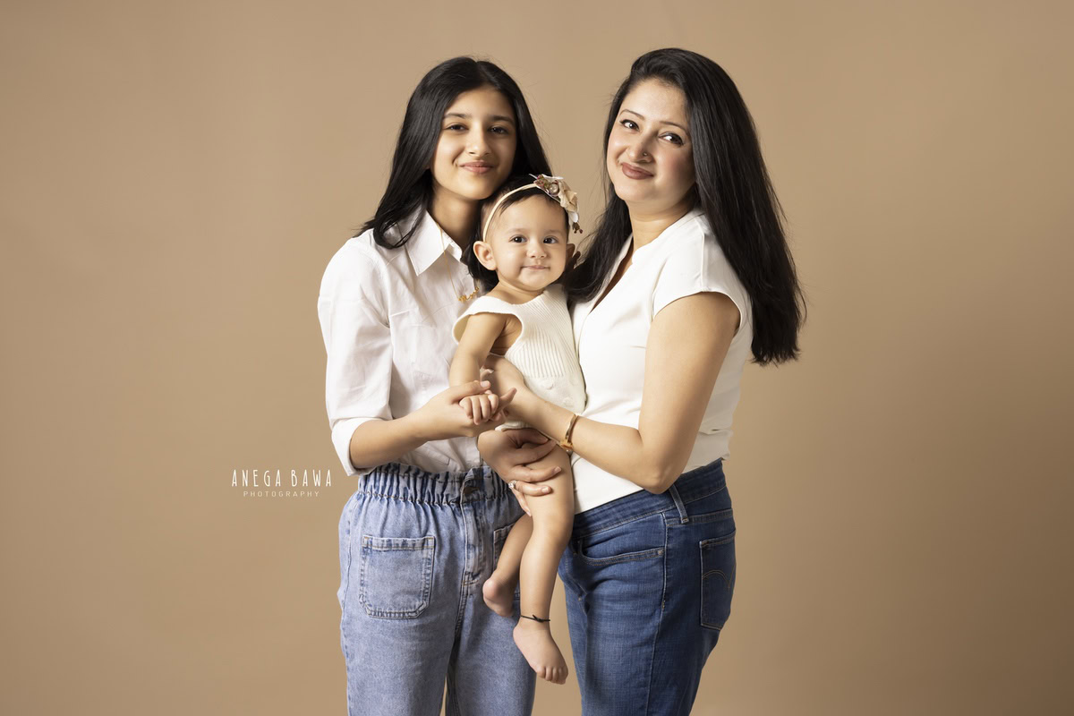 Mom holding her 10-month-old daughter, posing with her elder sister against a light brown backdrop. Captured by Anega Bawa Photography, Gurgaon (Delhi NCR).