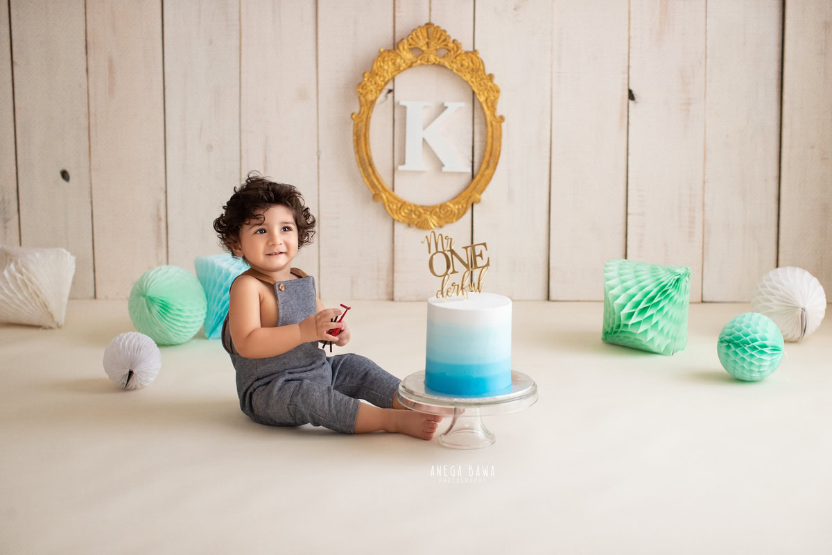 1 year old boy in blue dungaree, beige backdrop, cake smash, golden frame on wall. First birthday photo shoot in Delhi, Gurgaon. Captured by Anega Bawa family photographer Gurgaon (Delhi NCR).