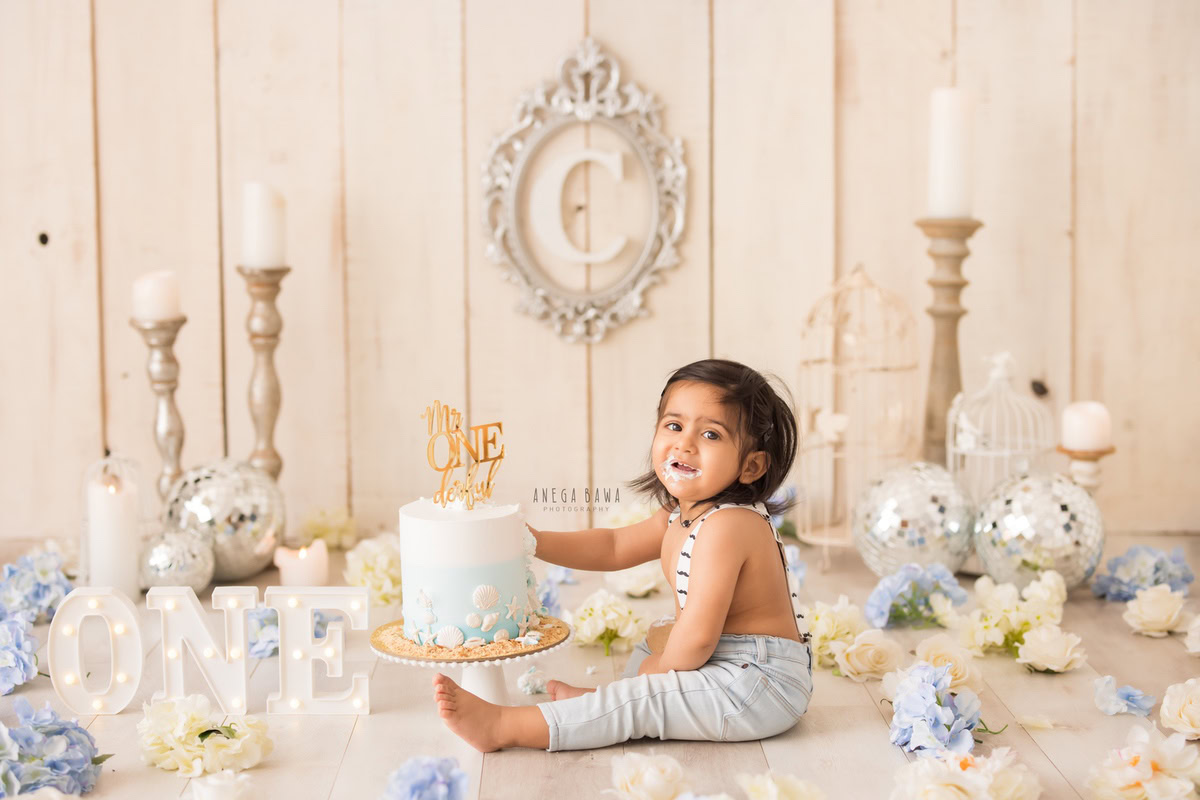 1 year old boy Cake Smash first birthday photoshoot in Delhi, Gurgaon with blue and ivory flowers, candle stand, disco balls, and silver alphabet frame on beige backdrop. Captured by Anega Bawa family photographer Gurgaon (Delhi NCR)