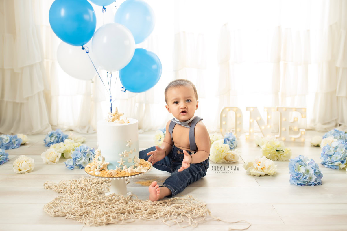 1year old boy with blue and white balloons, enjoying a cake smash session with a white backdrop, celebrating his first birthday. Cake Smash photo shoot in Delhi, Gurgaon by Anega Bawa Family Photographer Gurgaon (Delhi NCR).
