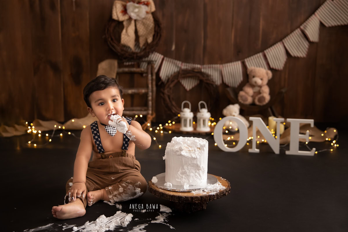 1 year boy in brown dungarees with fairy lights and teddy bears, enjoying a Cake Smash against a brown backdrop during a first birthday photoshoot by well-known Anega Bawa Photography in Delhi, Gurgaon.